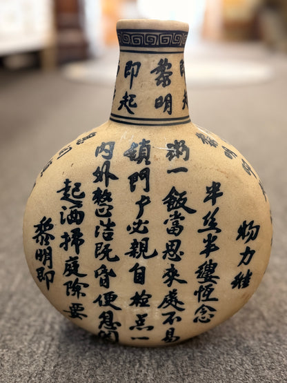 A round, flat-profile ceramic vase featuring black Asian calligraphy painted across a matte sand-colored surface.