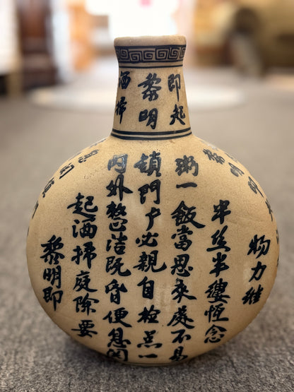 A round, flat-profile ceramic vase featuring black Asian calligraphy painted across a matte sand-colored surface.