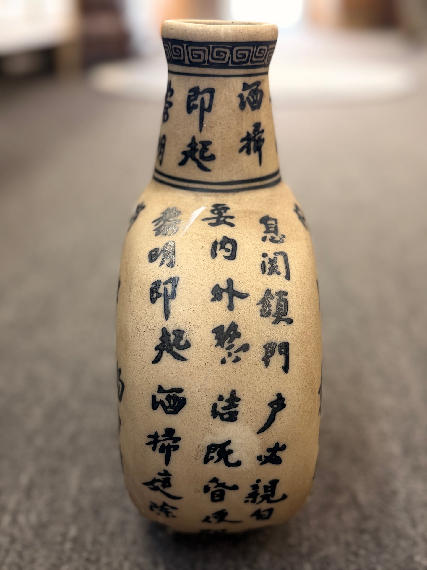 A round, flat-profile ceramic vase featuring black Asian calligraphy painted across a matte sand-colored surface.