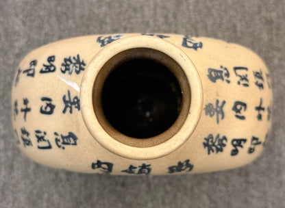 A round, flat-profile ceramic vase featuring black Asian calligraphy painted across a matte sand-colored surface.