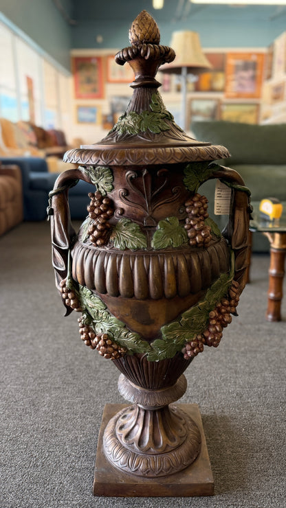 A large, ornate metal decorative floor urn with an antiqued bronze-style finish, painted high-relief green leaves and burgundy grape clusters, and a lid with an acorn finial.
