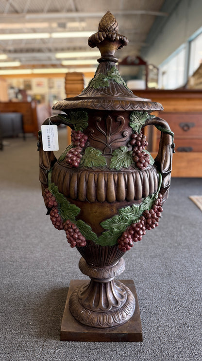A large, ornate metal decorative floor urn with an antiqued bronze-style finish, painted high-relief green leaves and burgundy grape clusters, and a lid with an acorn finial.