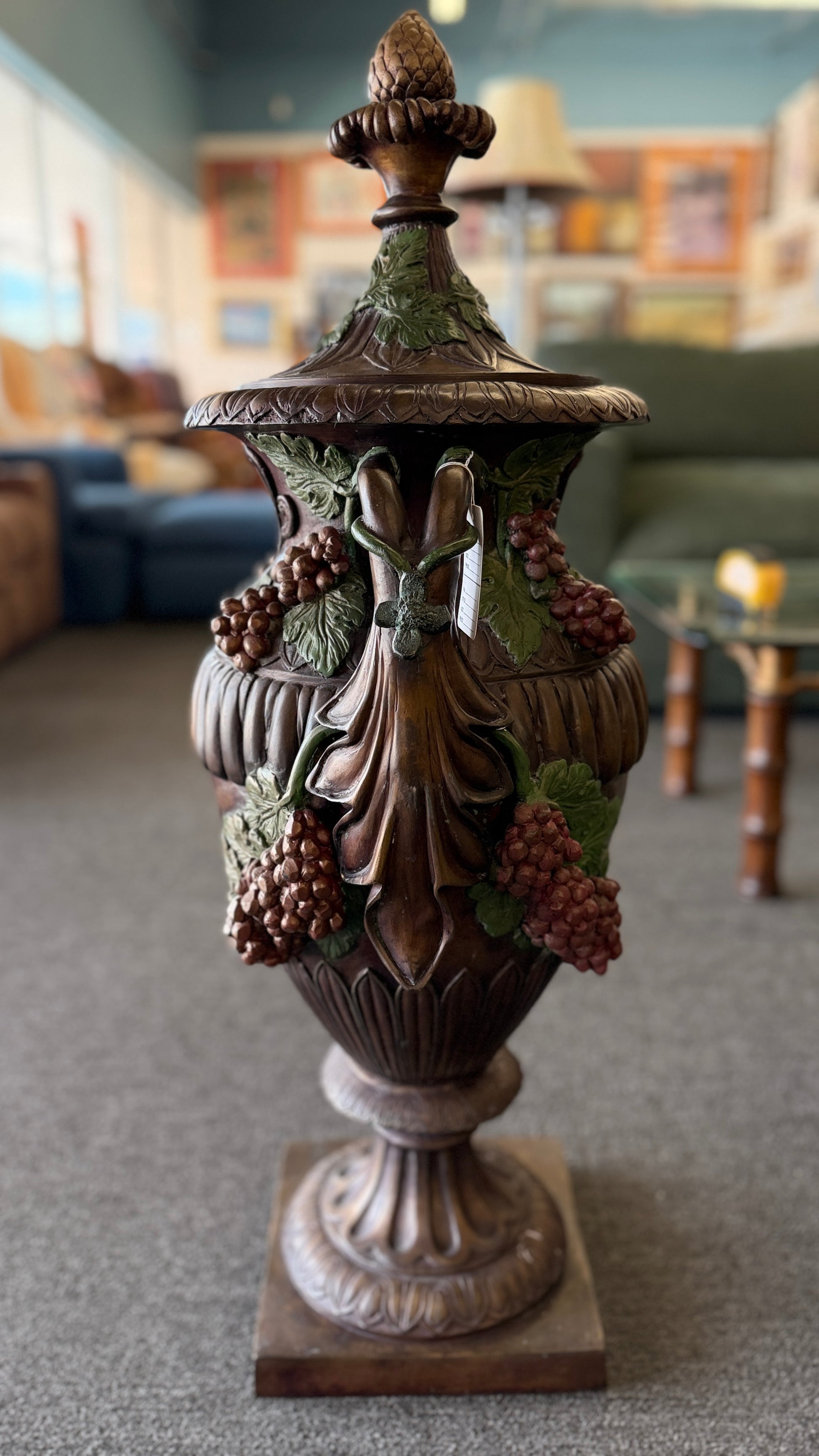 A large, ornate metal decorative floor urn with an antiqued bronze-style finish, painted high-relief green leaves and burgundy grape clusters, and a lid with an acorn finial.