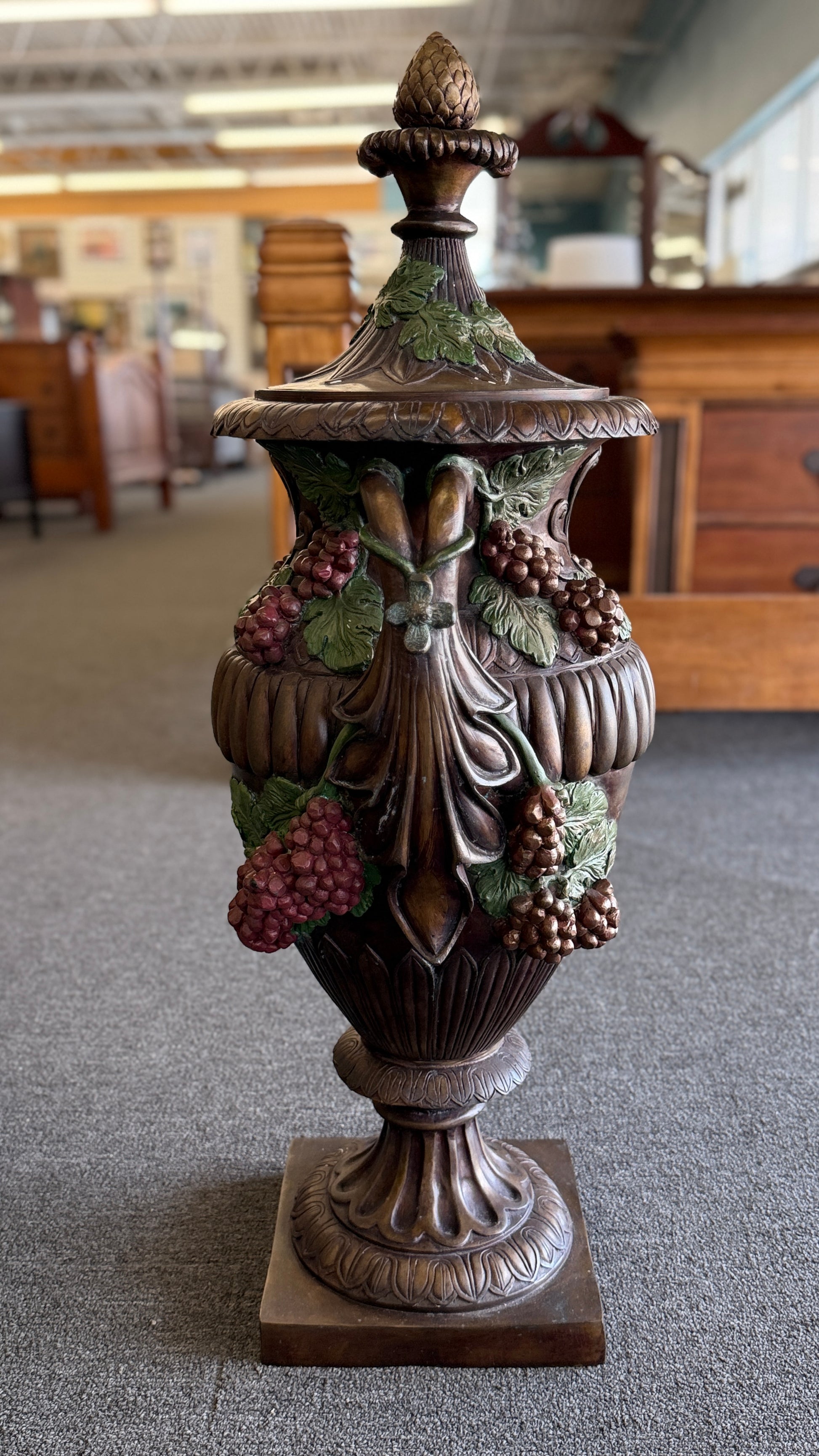 A large, ornate metal decorative floor urn with an antiqued bronze-style finish, painted high-relief green leaves and burgundy grape clusters, and a lid with an acorn finial.