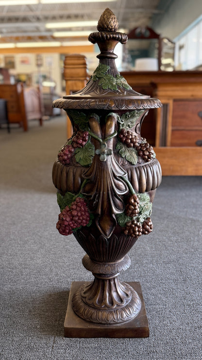 A large, ornate metal decorative floor urn with an antiqued bronze-style finish, painted high-relief green leaves and burgundy grape clusters, and a lid with an acorn finial.