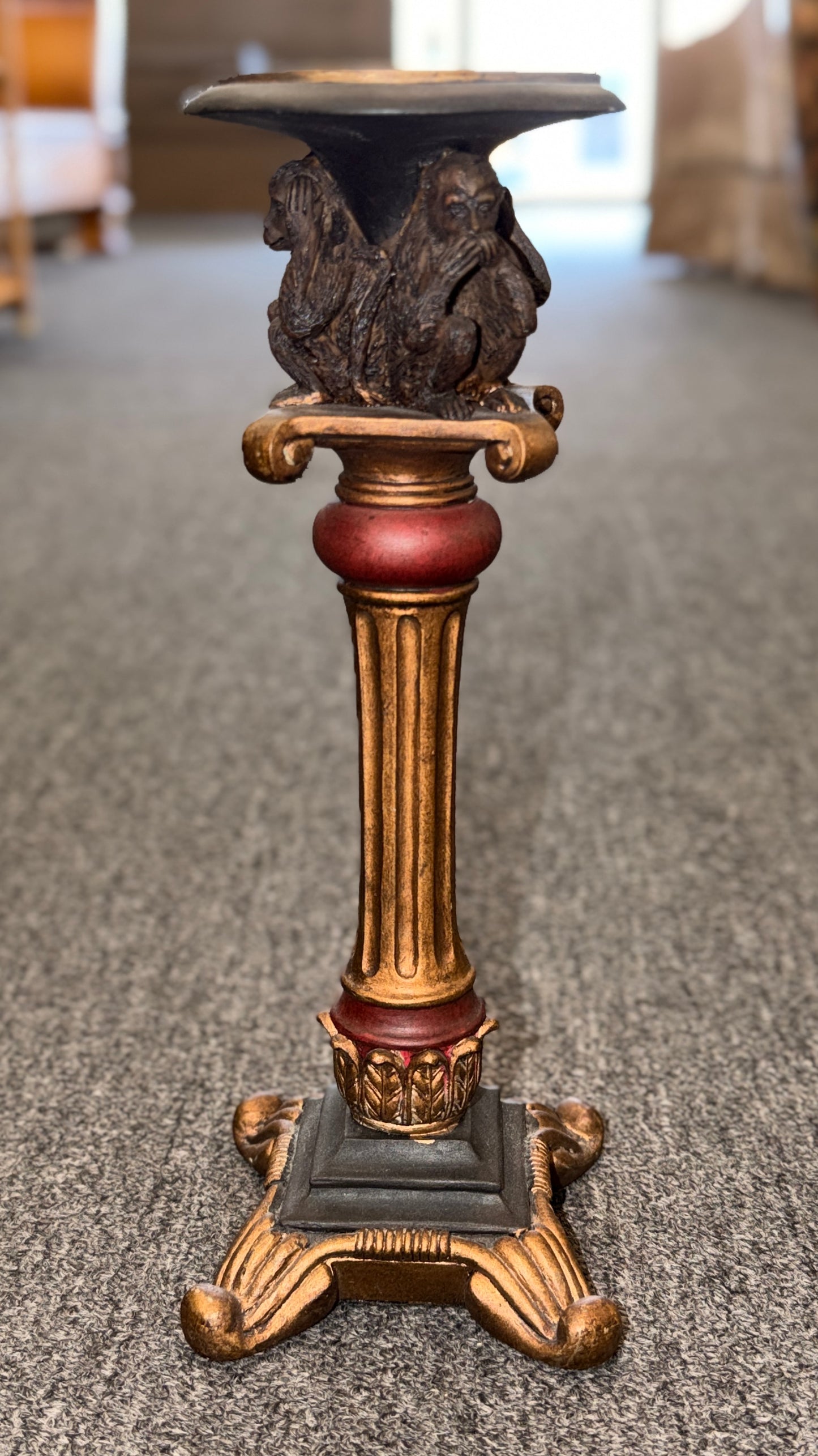 An ornate decorative ceramic candle holder with a fluted column base painted in antiqued gold and red tones, featuring a sculpted Three Wise Monkeys motif beneath the top tray.
