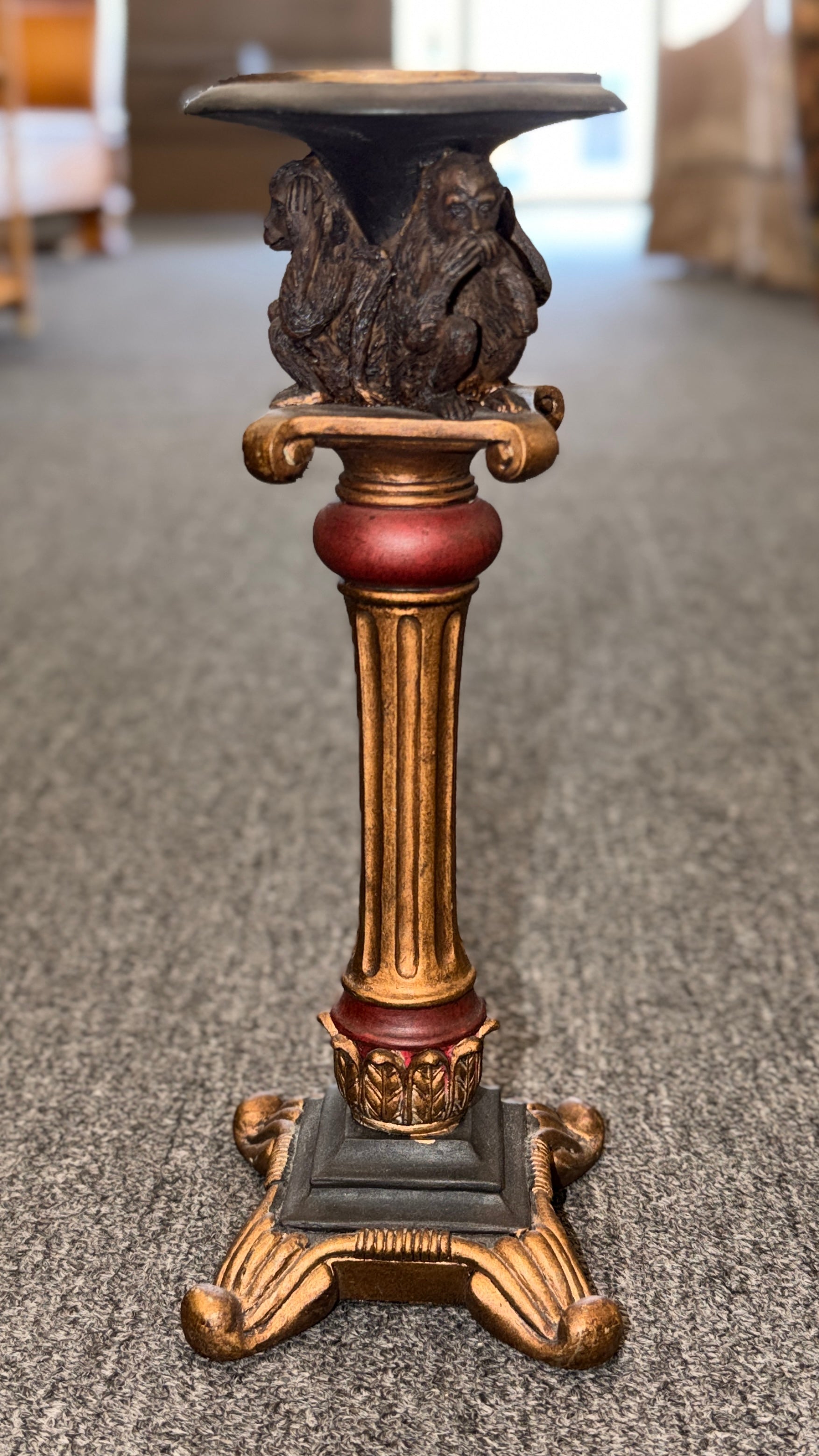 An ornate decorative ceramic candle holder with a fluted column base painted in antiqued gold and red tones, featuring a sculpted Three Wise Monkeys motif beneath the top tray.