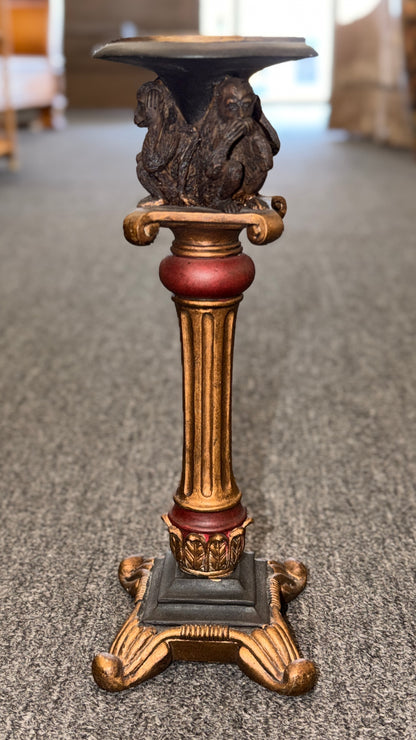 An ornate decorative ceramic candle holder with a fluted column base painted in antiqued gold and red tones, featuring a sculpted Three Wise Monkeys motif beneath the top tray.