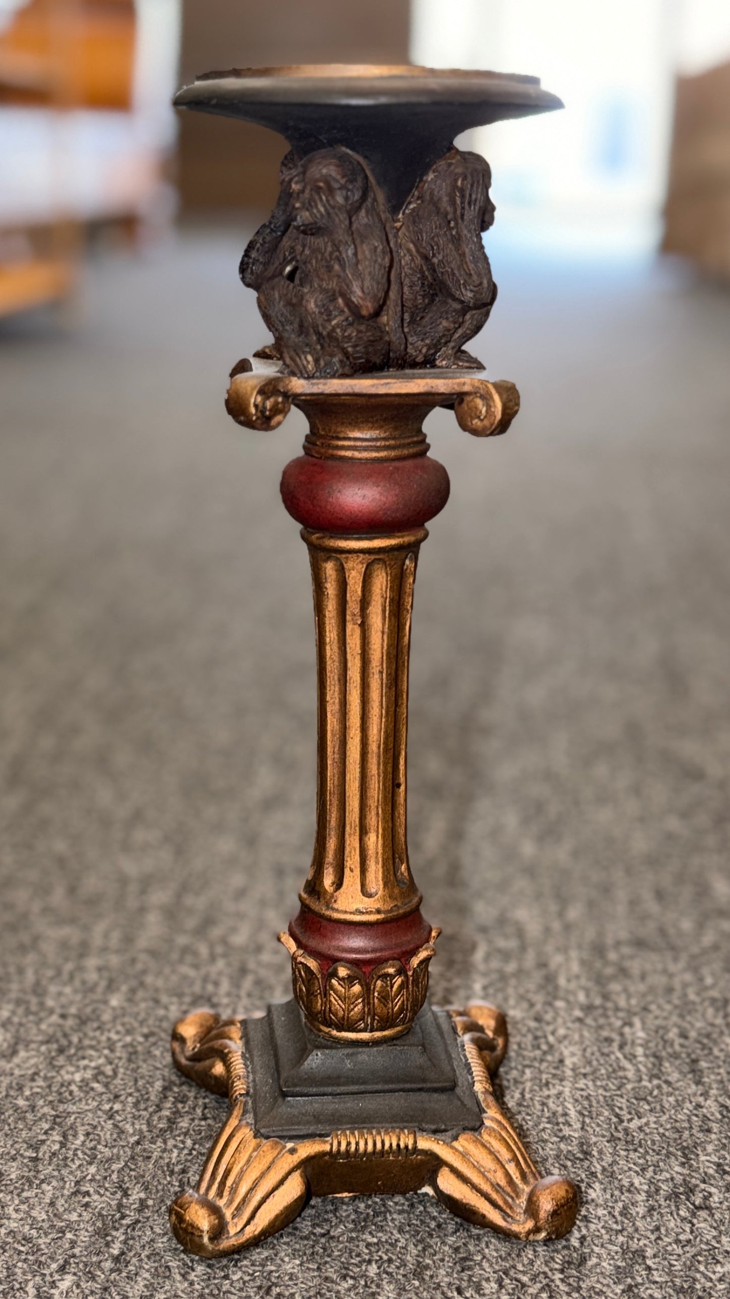 An ornate decorative ceramic candle holder with a fluted column base painted in antiqued gold and red tones, featuring a sculpted Three Wise Monkeys motif beneath the top tray.
