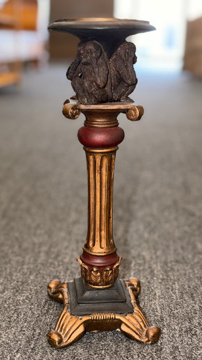 An ornate decorative ceramic candle holder with a fluted column base painted in antiqued gold and red tones, featuring a sculpted Three Wise Monkeys motif beneath the top tray.