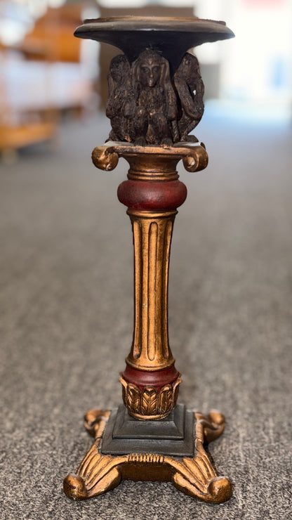An ornate decorative ceramic candle holder with a fluted column base painted in antiqued gold and red tones, featuring a sculpted Three Wise Monkeys motif beneath the top tray.