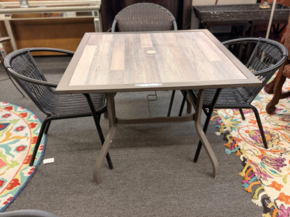 A 5-piece patio dining set featuring a square paneled top table with an umbrella hole and four dark woven resin wicker armchairs on iron frames.