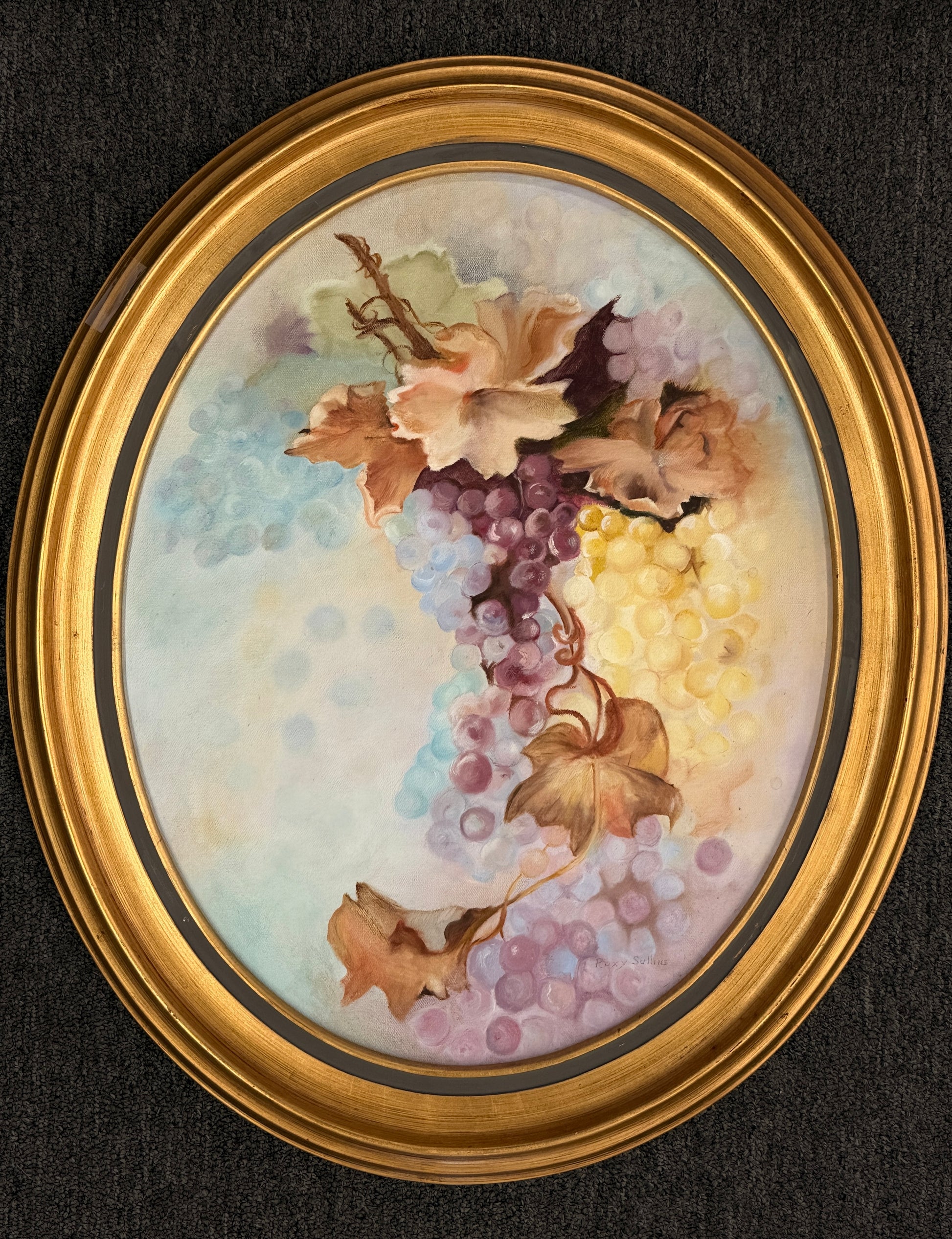 Oval framed still life artwork depicting a colorful cluster of grapes and leaves in soft pastel tones within a classic wooden frame.