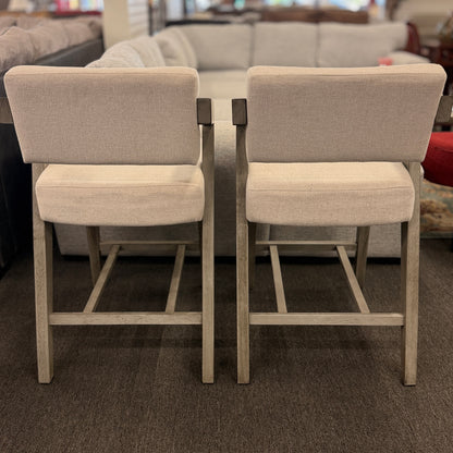 A pair of contemporary counter-height barstools featuring light oat upholstered seats and backs with angular weathered-wood frames in excellent condition.