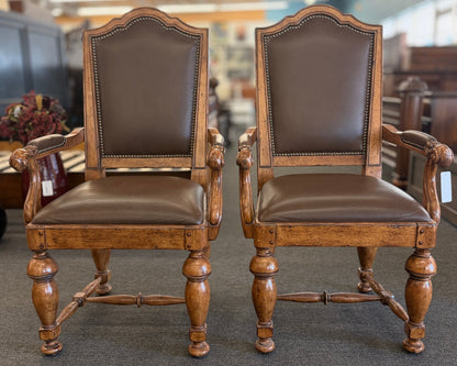Pair of traditional wooden armchairs featuring high arched backs, brown leather upholstery accented with nailhead trim, and turned legs with stretchers in excellent condition.