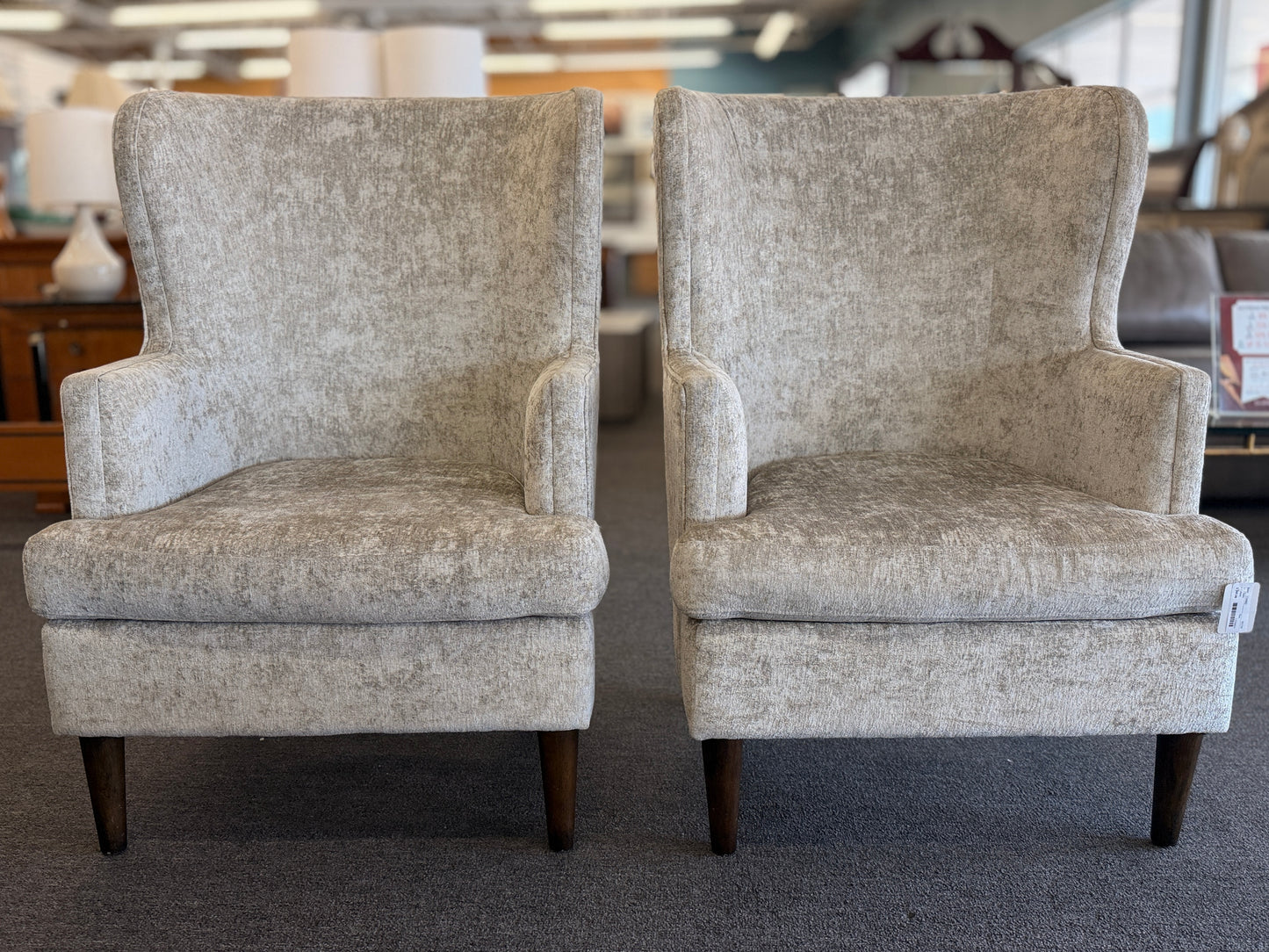 Matching pair of transitional wingback accent armchairs upholstered in textured light beige fabric with dark brown tapered wooden legs.