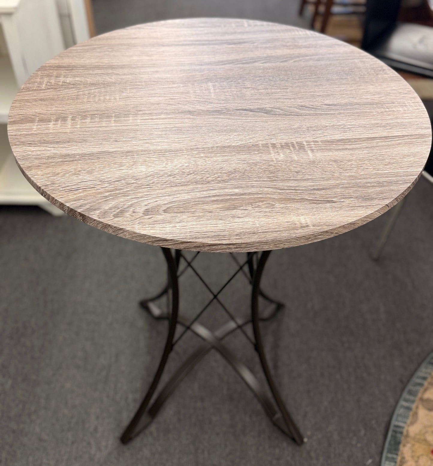 Round pub table with a light brownish-grey faux wood grain top and a dark metal pedestal base featuring curved intersecting legs.