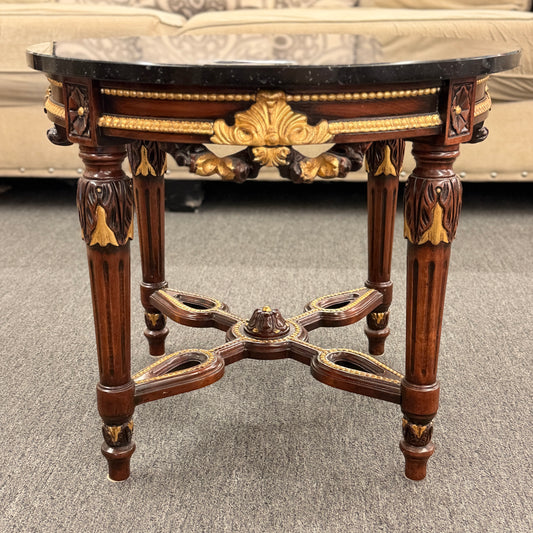 Round accent table featuring a dark marble top, ornate carved wooden base with gold-painted detailing, and a decorative cross-stretcher.