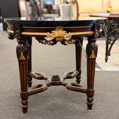 Round accent table featuring a dark marble top, ornate carved wooden base with gold-painted detailing, and a decorative cross-stretcher.