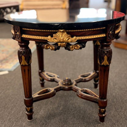 Round accent table featuring a dark marble top, ornate carved wooden base with gold-painted detailing, and a decorative cross-stretcher.