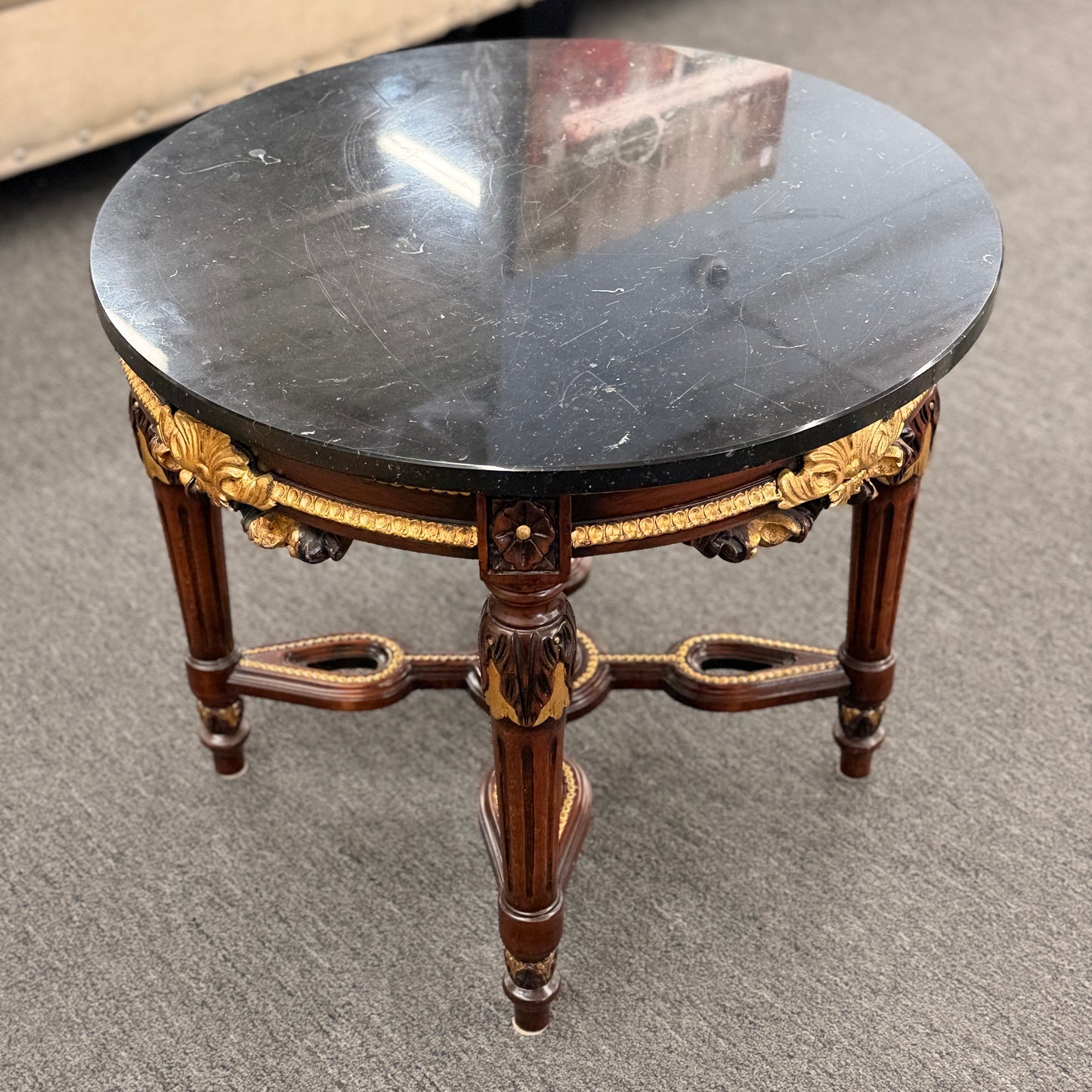 Round accent table featuring a dark marble top, ornate carved wooden base with gold-painted detailing, and a decorative cross-stretcher.