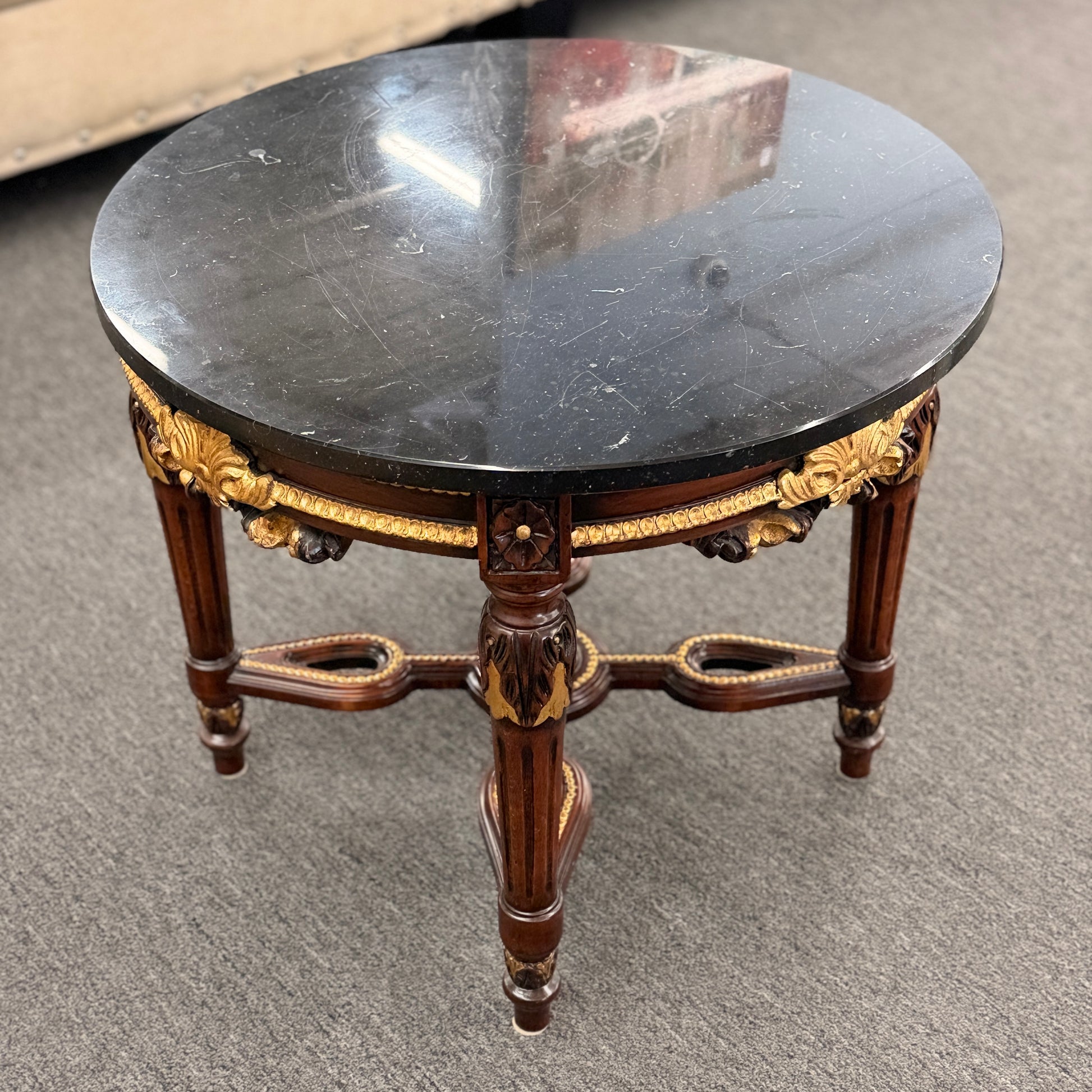 Round accent table featuring a dark marble top, ornate carved wooden base with gold-painted detailing, and a decorative cross-stretcher.