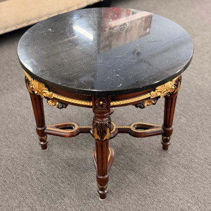 Round accent table featuring a dark marble top, ornate carved wooden base with gold-painted detailing, and a decorative cross-stretcher.