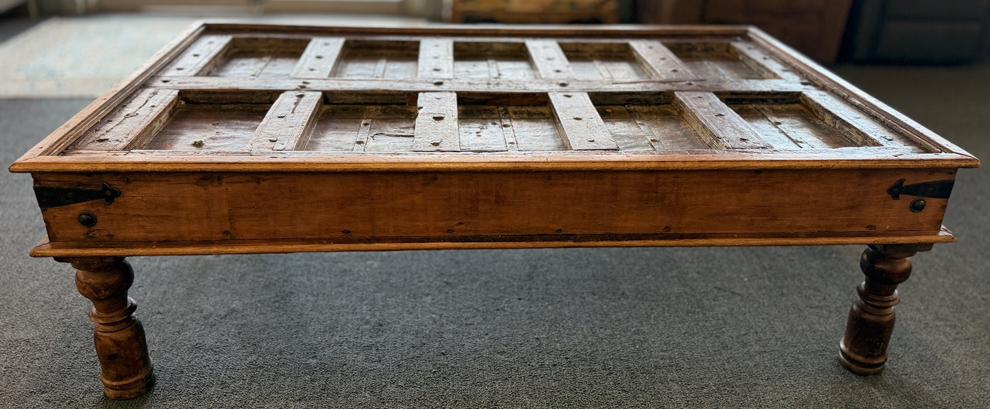 A rustic low-profile wooden coffee table featuring a paneled top resembling a repurposed antique door with decorative metal hinges and turned wooden legs.
