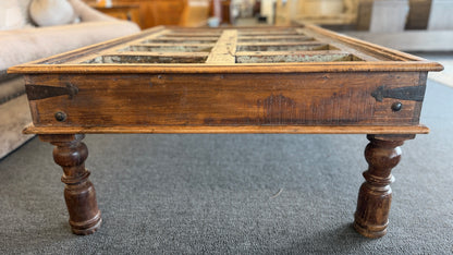 A rustic low-profile wooden coffee table featuring a paneled top resembling a repurposed antique door with decorative metal hinges and turned wooden legs.