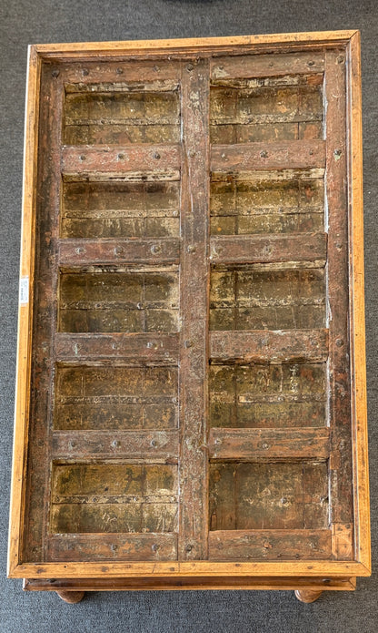A rustic low-profile wooden coffee table featuring a paneled top resembling a repurposed antique door with decorative metal hinges and turned wooden legs.