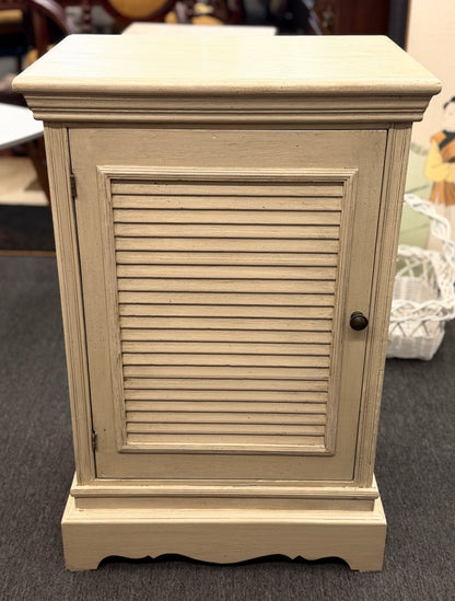 A small rustic accent cabinet with a distressed beige-cream painted finish, featuring a single louvered shutter door with a small dark round knob.