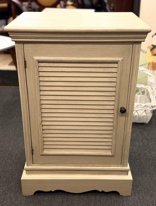 A small rustic accent cabinet with a distressed beige-cream painted finish, featuring a single louvered shutter door with a small dark round knob.
