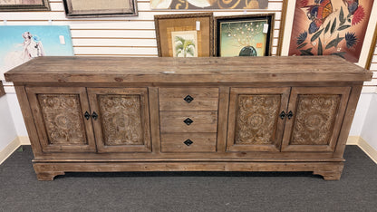 A large rustic Mediterranean-style credenza sideboard crafted from weathered wood featuring four relief-carved cabinet doors and three center drawers with dark ring pulls in good condition.