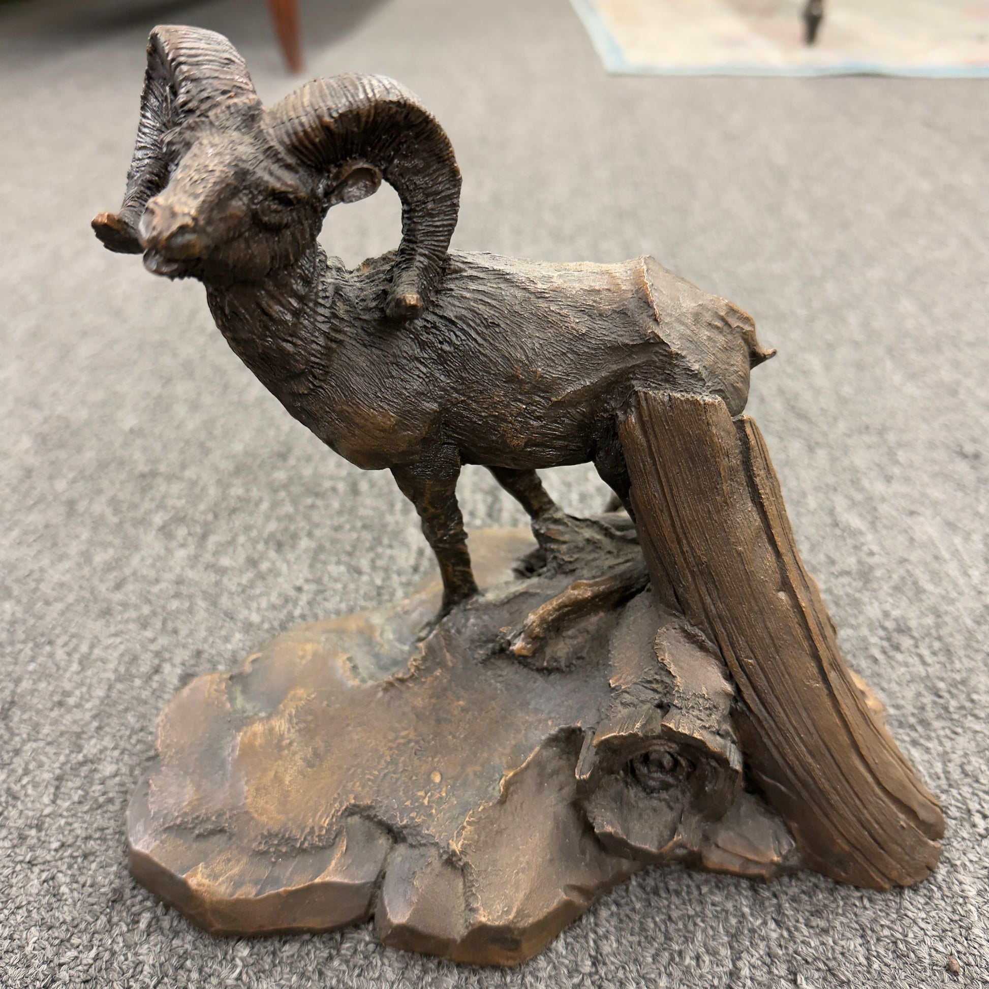 A rustic figurine depicting a ram standing on a rocky outcrop next to a broken tree stump.