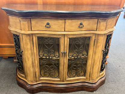 A rustic traditional wooden server featuring a curved silhouette, two top drawers, a dark top, and two glass doors with ornate metal scrollwork.