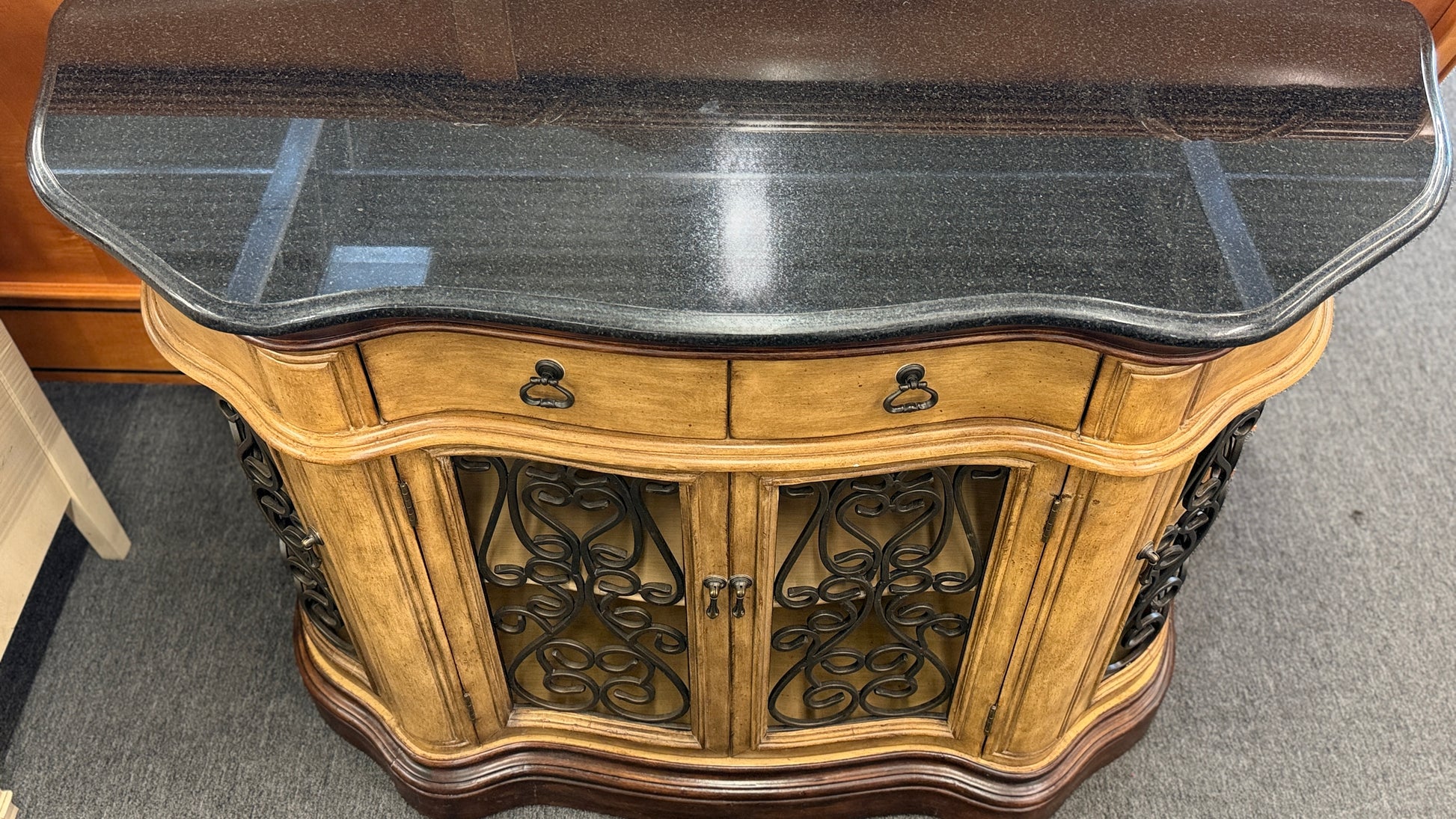 A rustic traditional wooden server featuring a curved silhouette, two top drawers, a dark top, and two glass doors with ornate metal scrollwork.