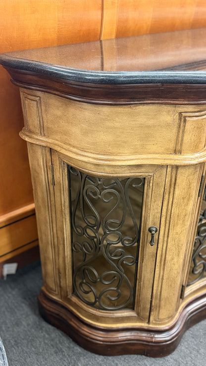 A rustic traditional wooden server featuring a curved silhouette, two top drawers, a dark top, and two glass doors with ornate metal scrollwork.