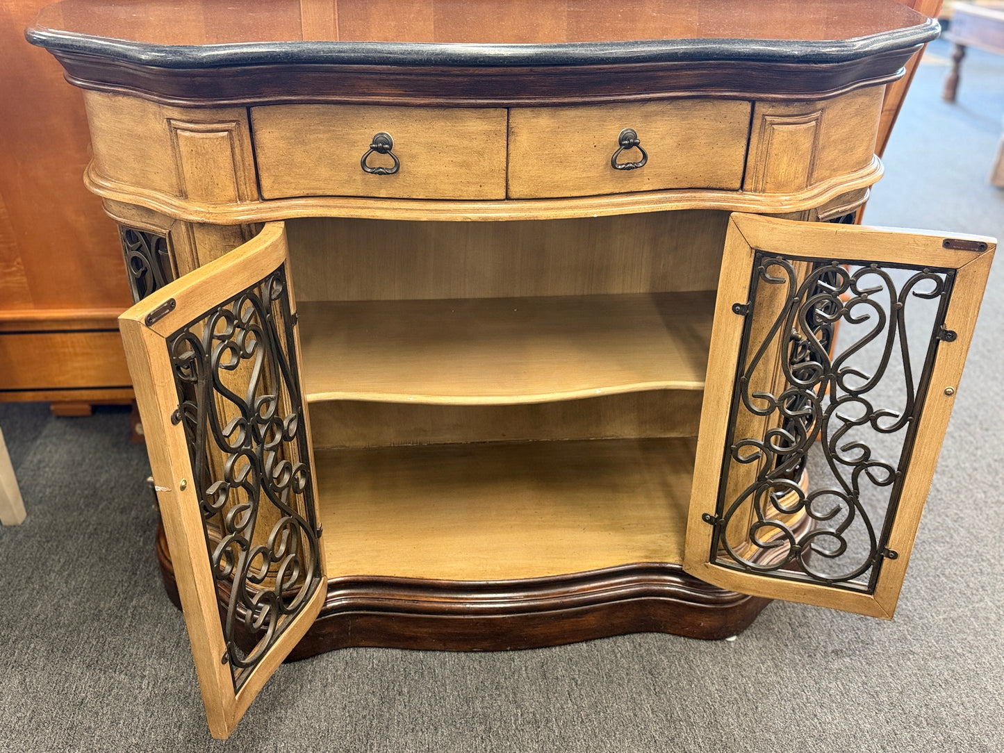A rustic traditional wooden server featuring a curved silhouette, two top drawers, a dark top, and two glass doors with ornate metal scrollwork.