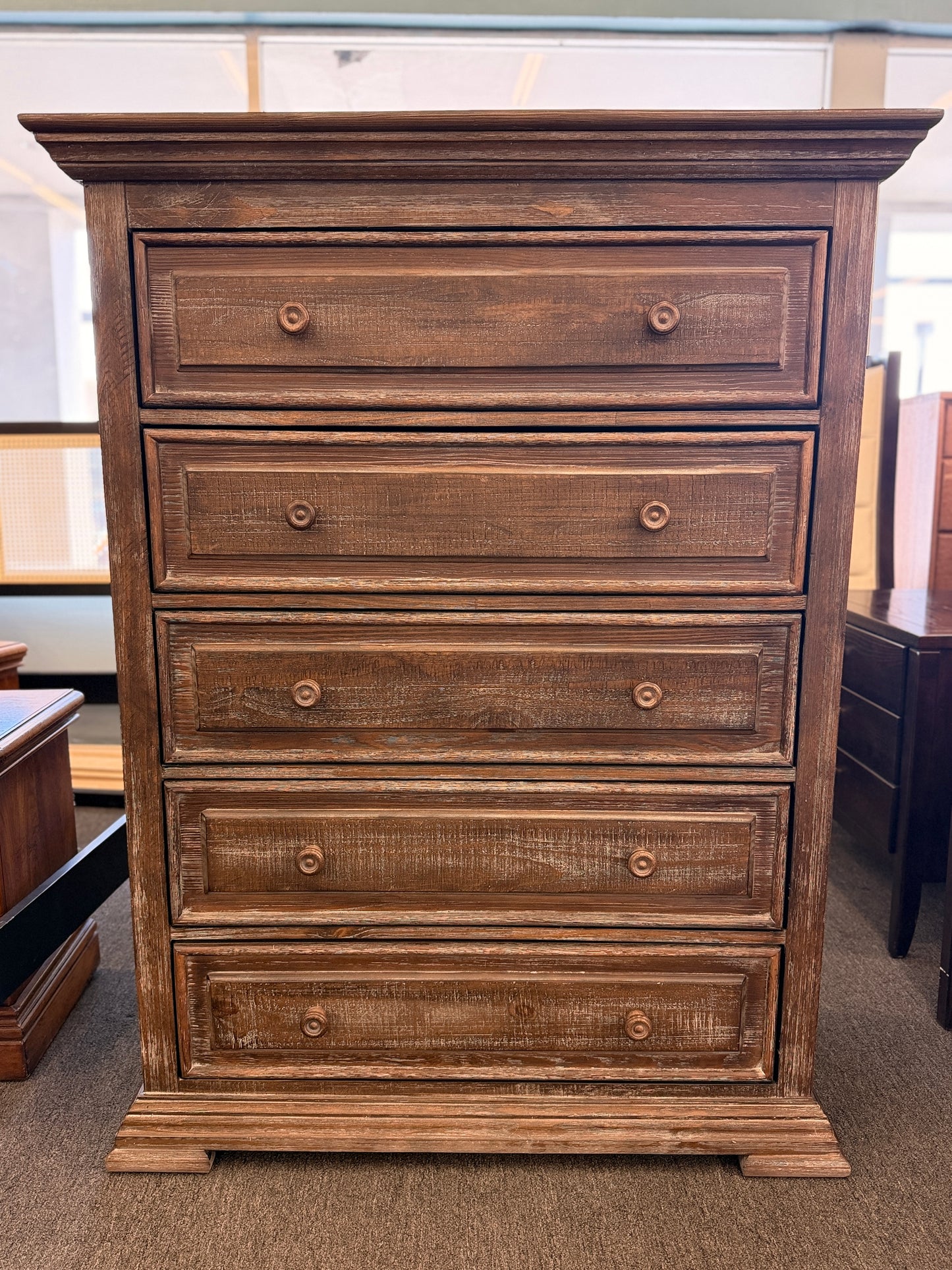 A tall rustic 5-drawer chest of drawers featuring a weathered white-washed finish over dark wood, a crown-molding top, and pairs of round matching knobs.
