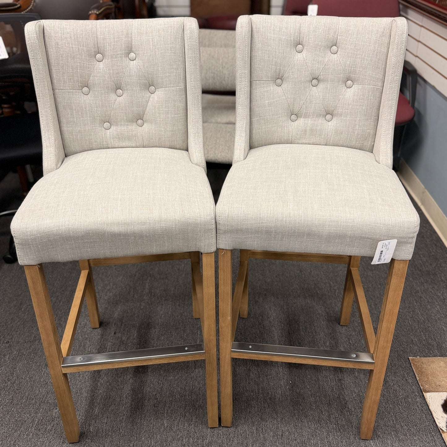 Set of two counter-height bar stools upholstered in textured beige fabric with diamond button-tufted backs, light wood legs, and metal footrest guards.