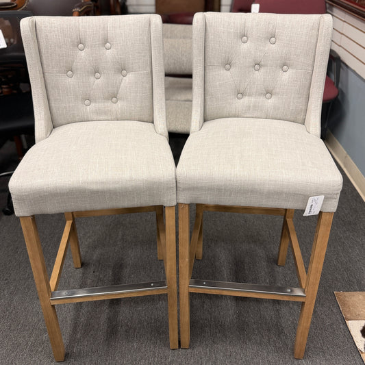 Set of two counter-height bar stools upholstered in textured beige fabric with diamond button-tufted backs, light wood legs, and metal footrest guards.