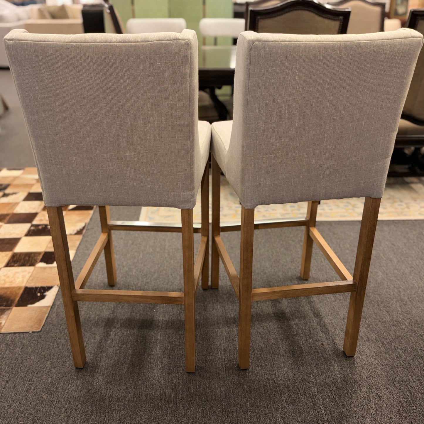 Set of two counter-height bar stools upholstered in textured beige fabric with diamond button-tufted backs, light wood legs, and metal footrest guards.
