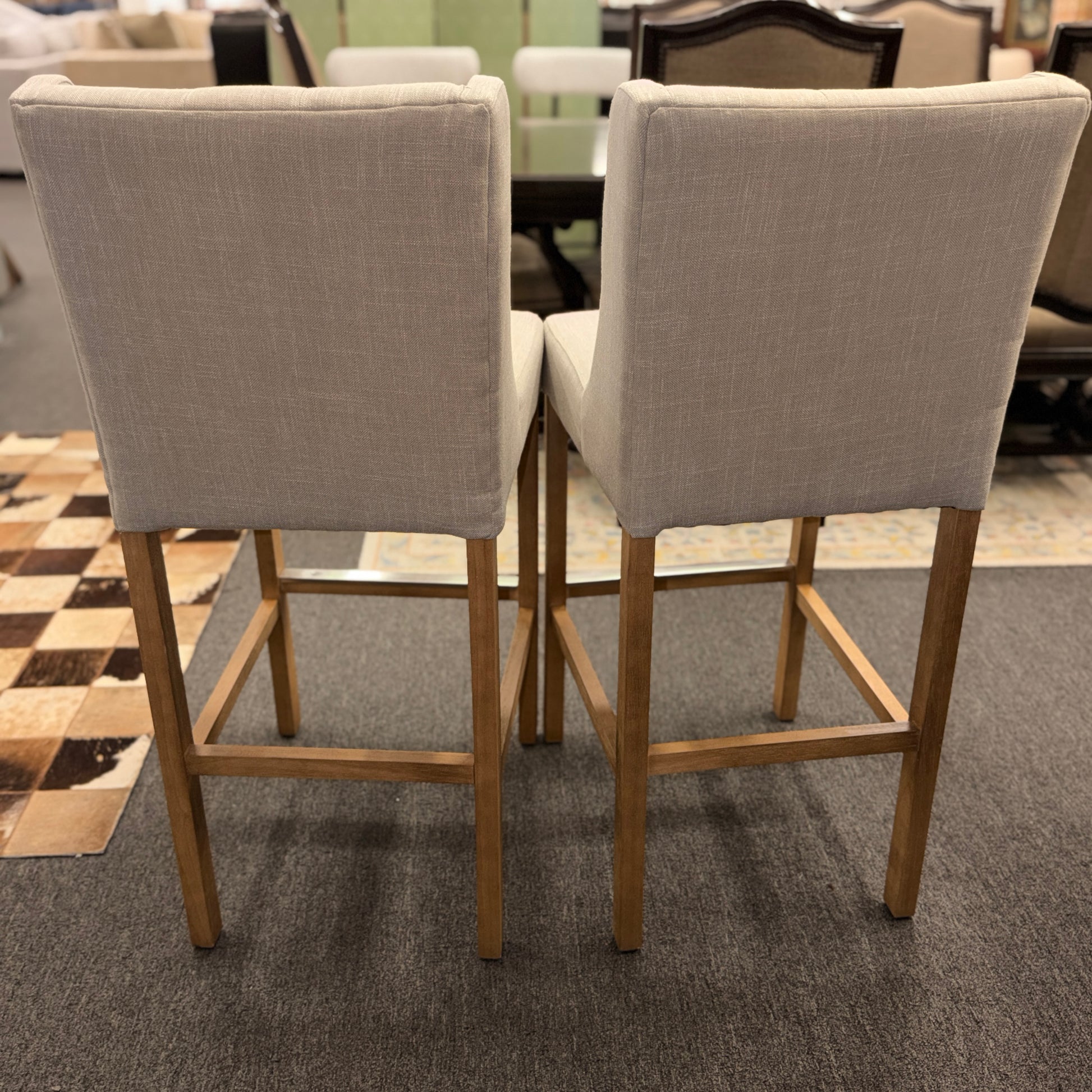 Set of two counter-height bar stools upholstered in textured beige fabric with diamond button-tufted backs, light wood legs, and metal footrest guards.
