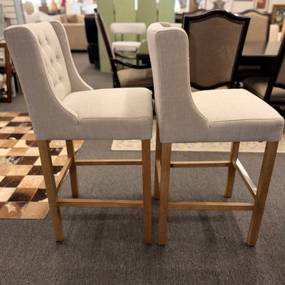 Set of two counter-height bar stools upholstered in textured beige fabric with diamond button-tufted backs, light wood legs, and metal footrest guards.