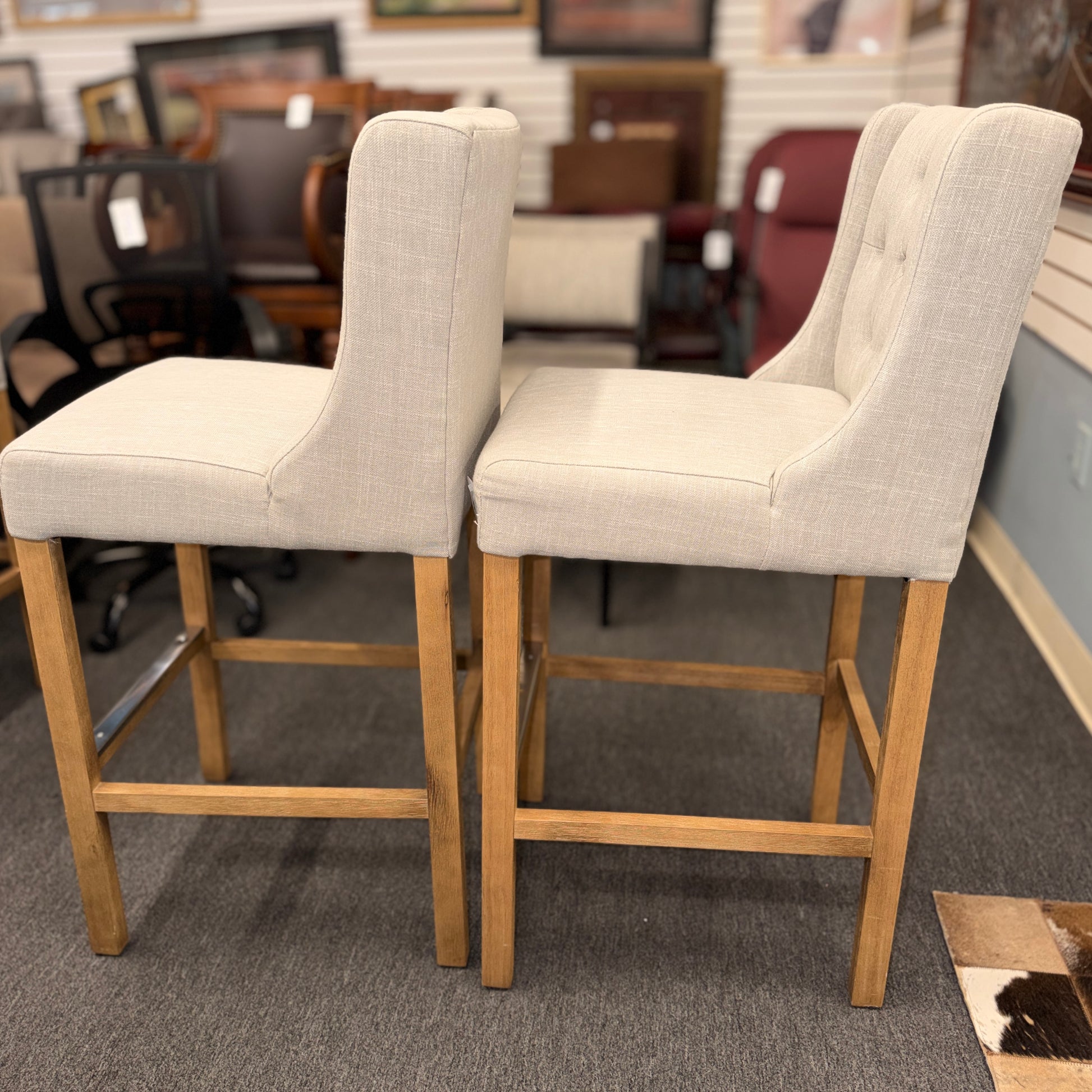 Set of two counter-height bar stools upholstered in textured beige fabric with diamond button-tufted backs, light wood legs, and metal footrest guards.