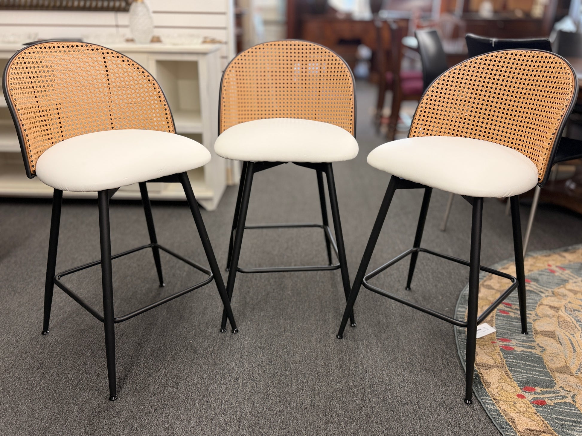 Set of three counter-height bar stools featuring curved woven cane backrests, white upholstered seat cushions, and black metal legs with built-in footrests.
