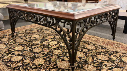 Square coffee table featuring a clear glass top insert, thick wooden frame, and an ornate scrolled metal base with traditional styling.