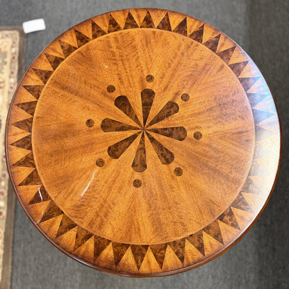 A round wooden pedestal side table featuring a geometric starburst inlay top, carved pineapple pedestal column, and tripod feet in good condition.