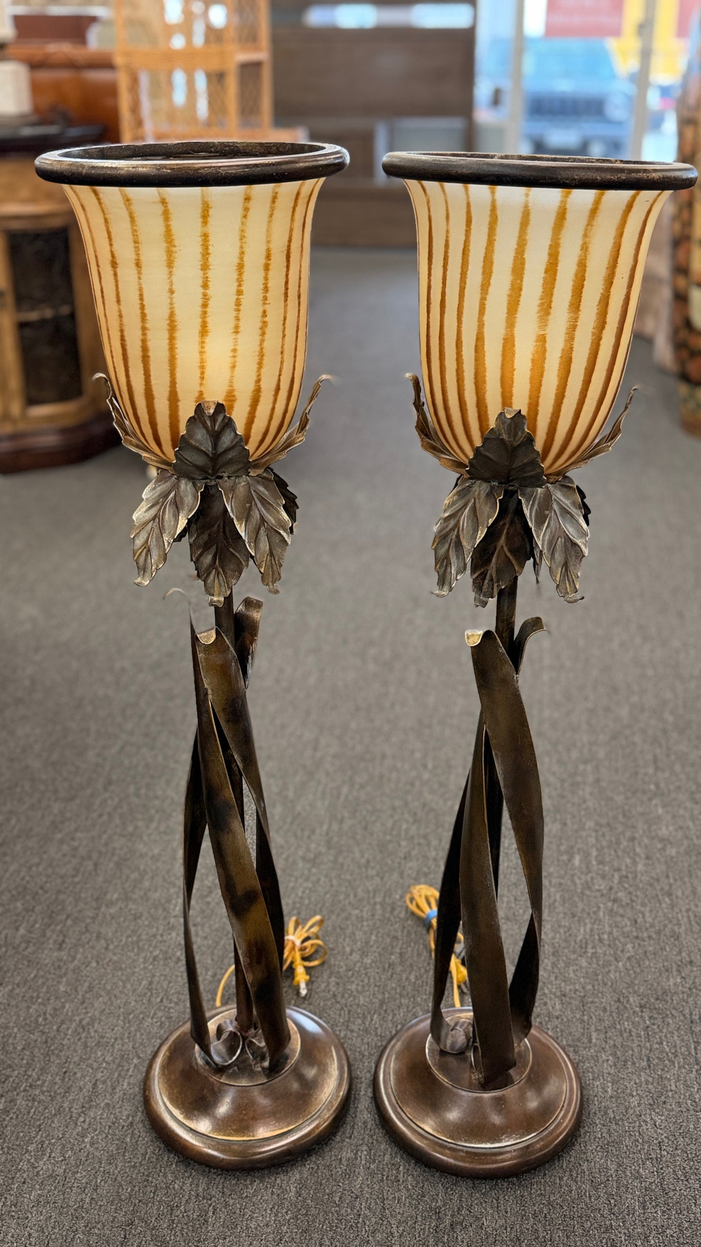 A pair of tall decorative table lamps featuring flared amber striped glass shades set in metal bases with sculpted leaves and twisted dark metal stems.