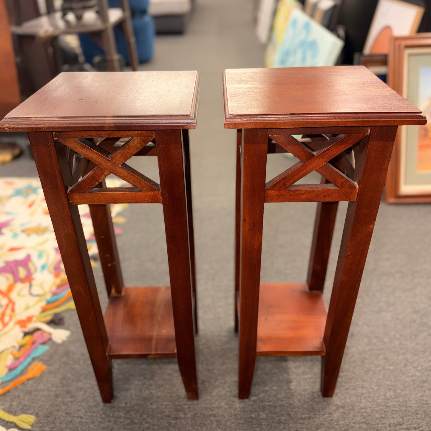 A pair of tall slim wooden side tables featuring square tops, lower display shelves, and cross-brace side details.